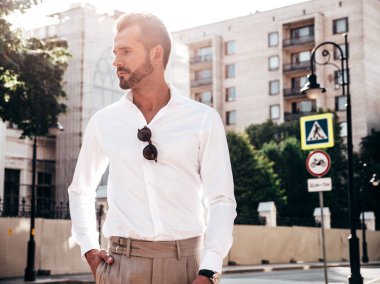 Yakışıklı, kendine güvenen, stil sahibi bir hipster Lambersexual modelin portresi. Beyaz gömlek ve pantolon giymiş modern bir adam. Moda erkeği gün batımında Avrupa şehrinin arka planında poz veriyor. Güneş gözlükleriyle