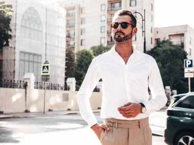 Yakışıklı, kendine güvenen, stil sahibi bir hipster Lambersexual modelin portresi. Beyaz gömlek ve pantolon giymiş modern bir adam. Moda erkeği gün batımında Avrupa şehrinin arka planında poz veriyor. Güneş gözlükleriyle