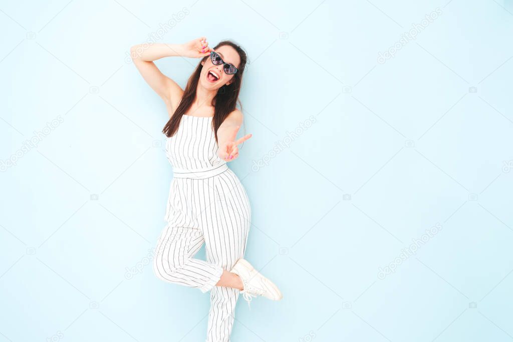 Portrait of young beautiful smiling female in trendy summer hipster overalls clothes. Sexy carefree woman posing near light blue wall in studio. Positive model having fun indoors.Shows peace sign