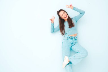 Yeni moda hippi kıyafetleri içinde gülümseyen güzel bir kadın. Stüdyoda açık mavi duvarın yanında poz veren kaygısız seksi kadın. Pozitif model içeride eğleniyor. Neşeli ve mutlu. Barış işareti nasıl?