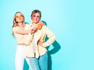 Moda kıyafetleri içinde gülümseyen iki güzel sarışın hippi kadın. Stüdyoda mavi duvarın yanında poz veren kaygısız seksi kadınlar. Moda ve pozitif modeller şekerleme lolipoplarıyla eğleniyorlar