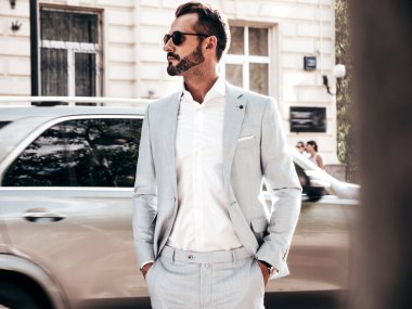 Yakışıklı, kendine güvenen, stil sahibi, lamberseksüel bir modelin portresi. Zarif beyaz takım elbise giymiş modern bir adam. Moda erkeği gün batımında Avrupa şehrinin arka planında poz veriyor. Güneş gözlükleriyle