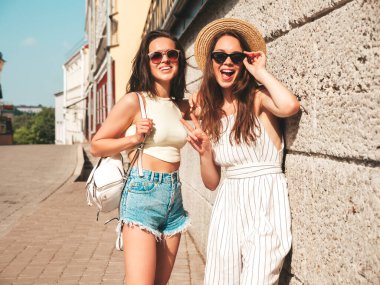 Son moda yaz kıyafetleri içinde iki güzel gülümseyen yeniyetme kadın. Sokakta şapkalı kayla poz veren kaygısız kadınlar. Pozitif saf modeller gün batımında eğleniyorlar, sarılıyorlar. 