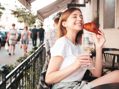 Genç, güzel, güler yüzlü, modaya uygun yaz kıyafetleri içinde bir bayan. Sokakta Veranda Kafe 'de poz veren kaygısız bir kadın. Beyaz şarap içen, tatilin tadını çıkaran, kruvasan yiyen bir kadın.