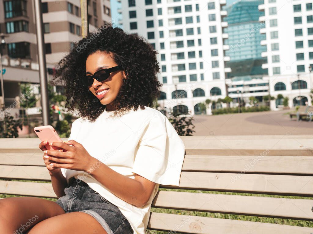 Beautiful black woman with afro curls hairstyle.Smiling model in white hoodie. Sexy carefree female posing on the street background in sunglasses. Looking at smartphone screen, using apps