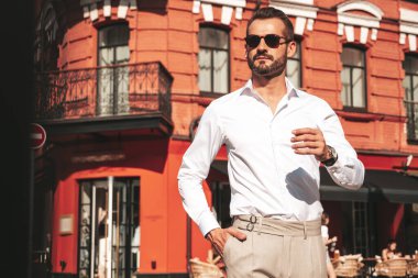 Yakışıklı, kendine güvenen, stil sahibi bir hipster Lambersexual modelin portresi. Beyaz gömlek ve pantolon giymiş modern bir adam. Moda erkeği gün batımında Avrupa şehrinin arka planında poz veriyor. Güneş gözlükleriyle