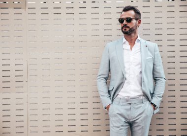 Yakışıklı, kendine güvenen, stil sahibi, lamberseksüel bir modelin portresi. Zarif beyaz takım elbise giymiş modern bir adam. Moda erkeği gün batımında çitlerin yanında poz veriyor. Güneş gözlükleriyle