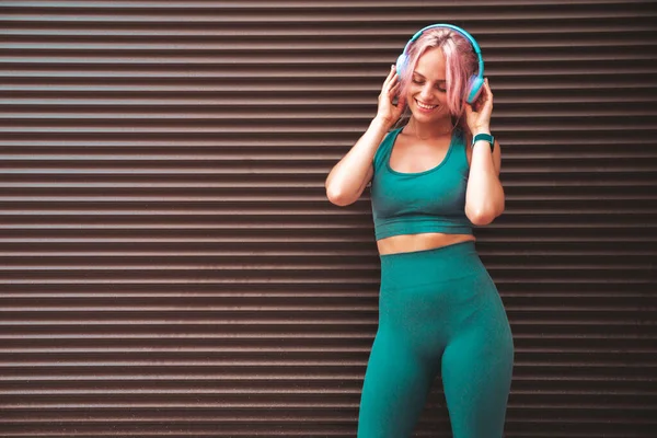 Fitness smiling woman in green sports clothing with pink hair. Young beautiful model with perfect body.Female in the street near roller shutter wall.Listening music in wireless headphones