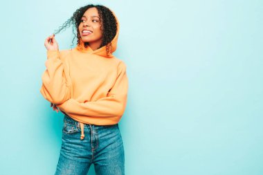 Afro kıvırcık saçlı, turuncu kapüşonlu ve kotlu güzel siyah kadın. Stüdyoda mavi duvarın yanında poz veren kaygısız seksi kadın. Neşeli ve mutlu