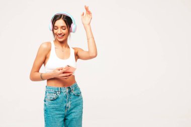 Kot pantolon giymiş, gülümseyen güzel bir kadın. Kablosuz kulaklıkla müzik dinleyen seksi tasasız model. Stüdyoda gri arka planda poz veren sevimli ve pozitif bir kadın. Akıllı telefon tutuluyor