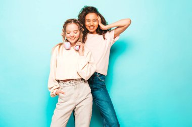 Moda kıyafetleri içinde iki güzel, gülümseyen uluslararası hippi kadın. Stüdyoda mavi duvarın yanında poz veren kaygısız seksi kadınlar. Pozitif modeller eğleniyor. Arkadaşlık kavramı