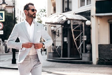 Yakışıklı, kendine güvenen, stil sahibi, lamberseksüel bir modelin portresi. Zarif beyaz takım elbise giymiş modern bir adam. Moda erkeği gün batımında Avrupa şehrinin arka planında poz veriyor. Güneş gözlükleriyle