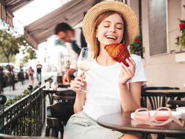 Genç, güzel, güler yüzlü, modaya uygun yaz kıyafetleri içinde bir bayan. Sokakta Veranda Kafe 'de poz veren kaygısız bir kadın. Beyaz şarap içen, tatilin tadını çıkaran, kruvasan yiyen bir kadın.