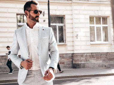 Yakışıklı, kendine güvenen, stil sahibi, lamberseksüel bir modelin portresi. Zarif beyaz takım elbise giymiş modern bir adam. Moda erkeği gün batımında Avrupa şehrinin arka planında poz veriyor. Güneş gözlükleriyle