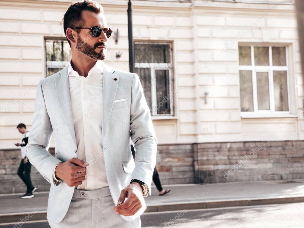 Portrait of handsome confident stylish hipster lambersexual model.Modern man dressed in elegant white suit. Fashion male posing in the street background in Europe city at sunset. In sunglasses