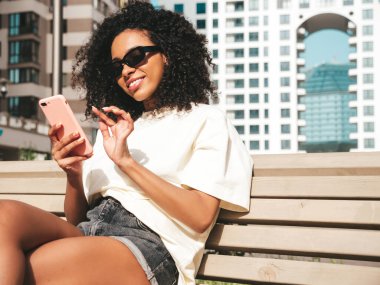 Saçları kıvırcık, güzel siyah kadın. Beyaz kapüşonlu, gülümseyen model. Sokakta güneş gözlükleriyle poz veren kaygısız seksi kadın. Akıllı telefon ekranına bakıyor, uygulamalar kullanıyor