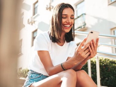 Güzel gülümseyen hippi bir kadının son moda kot şortlu portresi. Sokakta poz veren kaygısız seksi manken. Merdivenlerde oturuyor, akıllı telefon ekranına bakıyor, telefon uygulamalarını kullanıyor.