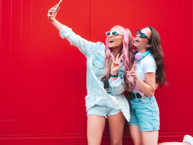 Son moda yaz kıyafetleri içinde iki güzel gülümseyen yeniyetme kadın. Sokakta pembe saçlı, kırmızı duvarın yanında poz veren kaygısız kadınlar. Pozitif saf modeller eğleniyor. Neşeli ve mutlu. Selfie çekiyor.