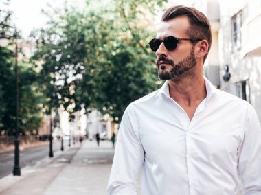 Yakışıklı, kendine güvenen, stil sahibi bir hipster Lambersexual modelin portresi. Beyaz gömlek ve pantolon giymiş modern bir adam. Moda erkeği gün batımında Avrupa şehrinin arka planında poz veriyor. Güneş gözlükleriyle