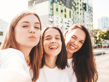 Üç güzel, gülümseyen yeniyetme kadın yazın moda olan aynı kıyafetleri giyiyor. Sokakta poz veren kaygısız seksi kadınlar. Pozitif modeller güneş gözlükleriyle eğleniyor. Selfie çekiyorlar.