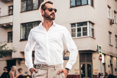 Yakışıklı, kendine güvenen, stil sahibi bir hipster Lambersexual modelin portresi. Beyaz gömlek ve pantolon giymiş modern bir adam. Moda erkeği gün batımında Avrupa şehrinin arka planında poz veriyor. Güneş gözlükleriyle