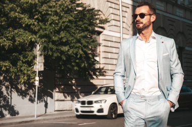 Yakışıklı, kendine güvenen, stil sahibi, lamberseksüel bir modelin portresi. Zarif beyaz takım elbise giymiş modern bir adam. Moda erkeği gün batımında Avrupa şehrinin arka planında poz veriyor. Güneş gözlükleriyle