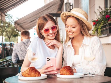 Son moda yaz kıyafetleri içinde iki güzel, gülümseyen yeniyetme kadın. Sokakta Veranda Kafe 'de oturan kaygısız kadınlar. Beyaz şarap içen, kruvasan yiyen, telefon ekranına bakan pozitif modeller.