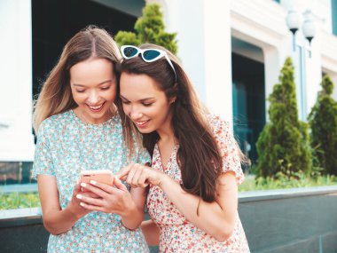 Son moda yaz kıyafetleri içinde iki güzel gülümseyen hippi kadın. Sokakta poz veren kaygısız kadınlar. Pozitif modeller eğleniyor, sarılıyorlar. Akıllı telefon ekranına bakıyorlar, telefon uygulamaları kullanıyorlar.