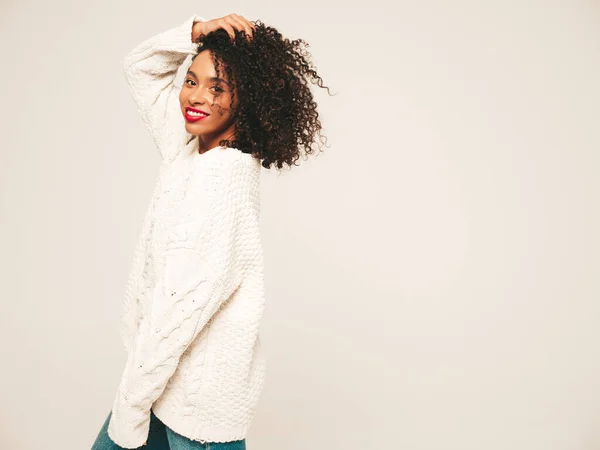 Beautiful black woman with afro curls hairstyle.Smiling model wearing warm white winter sweater and jeans clothes. Sexy carefree female posing on white background in studio. Happy and cheerful