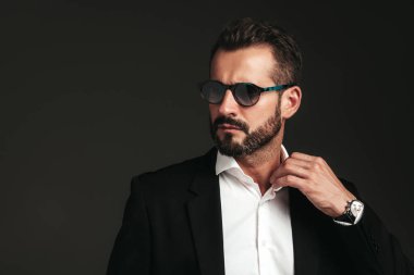 Yakışıklı, kendine güvenen, havalı hipster Lambersex modelinin yakın plan portresi. Zarif siyah takım elbiseli seksi modern adam. Moda erkeği karanlık arka planda stüdyoda poz veriyor. Güneş gözlükleriyle