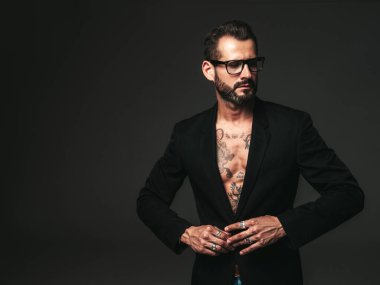 Yakışıklı, kendine güvenen, stil sahibi hipster Lambersex modelinin portresi. Siyah ceket giyen seksi modern adam. Dövmeli çıplak gövde. Stüdyoda siyah arka planda poz veren moda erkek. Gözlüklerle