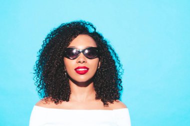 Saçları kıvırcık, güzel siyah bir kadın. Moda kıyafetleri içinde gülümseyen bir model. Stüdyoda mavi duvarın yanında poz veren kaygısız seksi kadın. Bronz tenli ve kırmızı dudaklı. Güneş gözlükleriyle