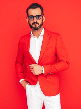 Yakışıklı, kendine güvenen, stil sahibi hipster Lambersex modelinin portresi. Zarif takım elbiseli seksi modern adam. Stüdyoda güneş gözlüklü, kırmızı duvarın yanında poz veren moda erkek.