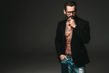 Yakışıklı, kendine güvenen, stil sahibi hipster Lambersex modelinin portresi. Siyah ceket giyen seksi modern adam. Dövmeli çıplak gövde. Stüdyoda siyah arka planda poz veren moda erkek. Gözlüklerle