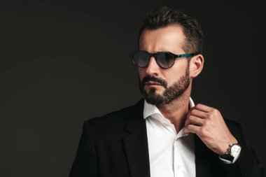 Yakışıklı, kendine güvenen, havalı hipster Lambersex modelinin yakın plan portresi. Zarif siyah takım elbiseli seksi modern adam. Moda erkeği karanlık arka planda stüdyoda poz veriyor. Güneş gözlükleriyle