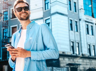 Yakışıklı, güler yüzlü, stil sahibi, lamberseksüel model. Mavi gömlekli, modern bir adam. Sokakta güneş gözlükleriyle poz veren modacı erkek. Gün batımında dışarıda. Akıllı telefon uygulamaları kullanıyor. SMS yazıyor.