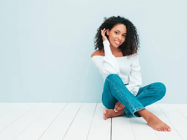 Beautiful black woman with afro curls hairstyle.Smiling model in sweater and trendy jeans clothes. Sexy carefree female sitting near light blue wall in studio. Tanned and cheerful