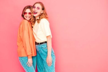 İki genç, güzel, gülümseyen yeniyetme kadın, modaya uygun yaz hippi kıyafetleri içinde. Stüdyoda pembe duvarın yanında poz veren kaygısız seksi kadınlar. Moda ve pozitif modeller eğleniyor. Neşeli ve mutlu