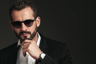 Yakışıklı, kendine güvenen, havalı hipster Lambersex modelinin yakın plan portresi. Zarif siyah takım elbiseli seksi modern adam. Moda erkeği karanlık arka planda stüdyoda poz veriyor. Güneş gözlükleriyle