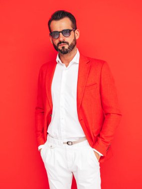 Yakışıklı, kendine güvenen, stil sahibi hipster Lambersex modelinin portresi. Zarif takım elbiseli seksi modern adam. Stüdyoda güneş gözlüklü, kırmızı duvarın yanında poz veren moda erkek.