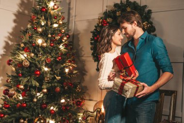 Gülümseyen güzel kadın ve yakışıklı erkek arkadaşı. Mutlu neşeli aile Noel ağacının yanında poz veriyor. Mankenler sarılıyor. Aşk, Noel konsepti. Birbirlerine hediye kutuları tutuyorlar.