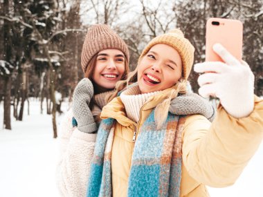 Son moda sıcak kıyafetler ve eşarplar içinde iki güzel, gülümseyen hippi kadın. Parktaki sokakta poz veren tasasız kadınlar. Pozitif saf modeller karda eğleniyor. Kış anlarının tadını çıkarıyorum. Selfie çekiyorum.