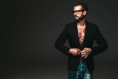 Yakışıklı, kendine güvenen, stil sahibi hipster Lambersex modelinin portresi. Siyah ceket giyen seksi modern adam. Dövmeli çıplak gövde. Stüdyoda siyah arka planda poz veren moda erkek. Gözlüklerle