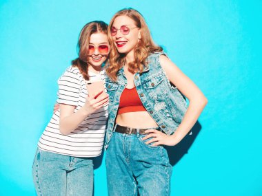Moda kıyafetleri içinde gülümseyen iki güzel hippi kadın. Stüdyoda mavi duvarın yanında poz veren kaygısız seksi kadınlar. Moda ve akıllı telefon ekranına bakan pozitif modeller. Telefon uygulamaları kullanarak.