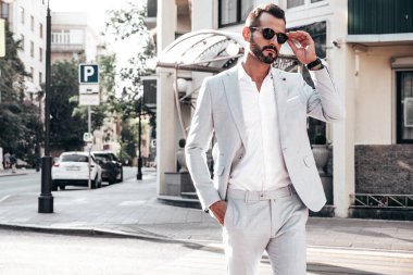 Yakışıklı, kendine güvenen, stil sahibi, lamberseksüel modelin portresi. Zarif beyaz takım elbise giymiş modern bir adam. Moda erkeği gün batımında Avrupa şehrinin arka planında poz veriyor. Güneş gözlükleriyle