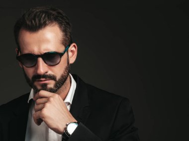 Yakışıklı, kendine güvenen, havalı hipster Lambersex modelinin yakın plan portresi. Zarif siyah takım elbiseli seksi modern adam. Moda erkeği karanlık arka planda stüdyoda poz veriyor. Güneş gözlüklü. Düşünceli. 