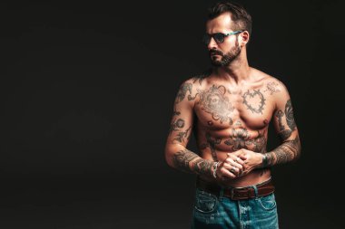 Yakışıklı, kendine güvenen, stil sahibi hipster Lambersex modelinin portresi. Seksi, modern adam. Dövmeli çıplak gövde. Stüdyoda siyah arka planda poz veren moda erkek. Güneş gözlükleriyle