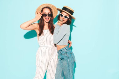 Son moda beyaz tişört ve kot pantolon giyen iki güzel, gülümseyen yeniyetme kadın stüdyoda açık mavi duvarın yanında poz veren kaygısız kadınlar. Şapkalı pozitif ve neşeli modeller. Dilini gösterir