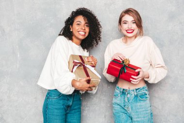 Gri duvarın yanında poz veren iki güzel, güler yüzlü uluslararası hippi kadın. Pozitif modeller birbirlerine hediye kutuları veriyorlar. Noel, Noel, Noel, Noel,