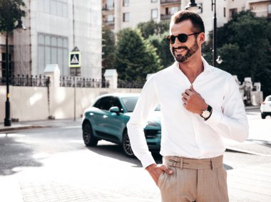 Yakışıklı, güler yüzlü, stil sahibi, lamberseksüel modelin portresi. Beyaz gömlek ve pantolon giymiş modern bir adam. Moda erkeği gün batımında Avrupa 'da sokak arkasındaymış gibi poz veriyor. Güneş gözlükleriyle